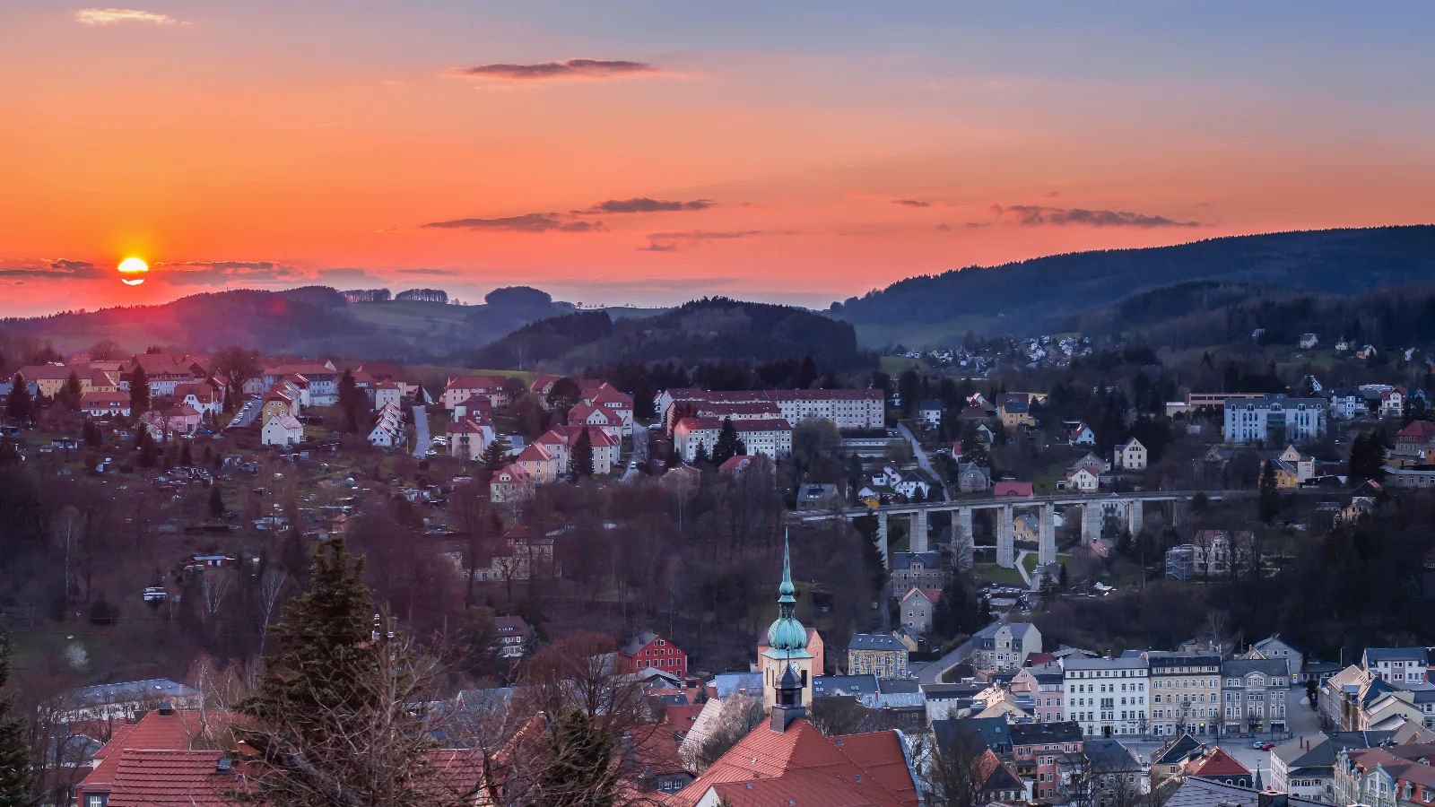 Sonnenuntergang über Sebnitz mit Blick auf die Stadt, die umliegenden Hügel und die beleuchteten Häuser im warmen Abendlicht